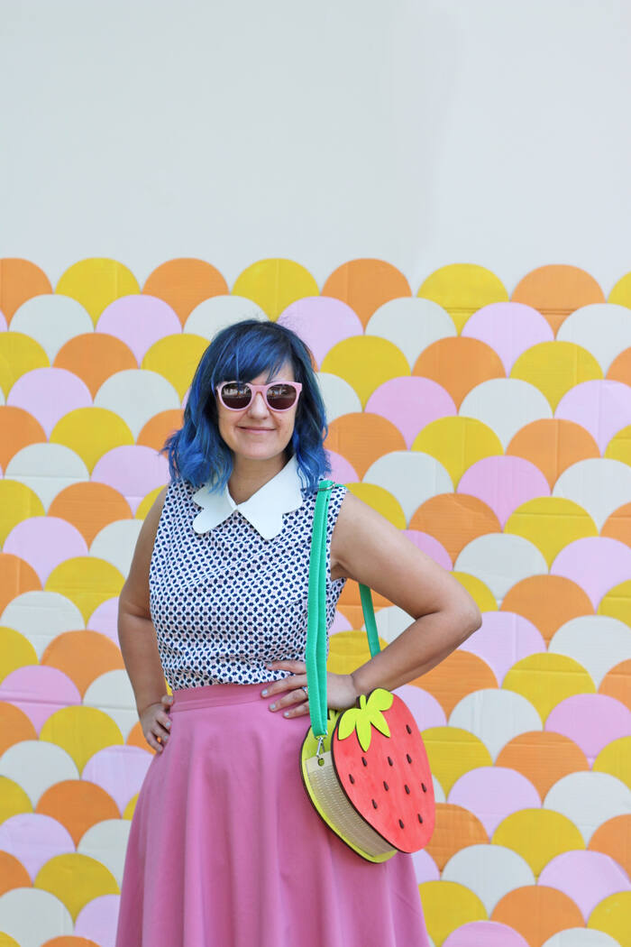 A woman with blue hair and a strawberry purse in front of a pink and orange scalloped wall