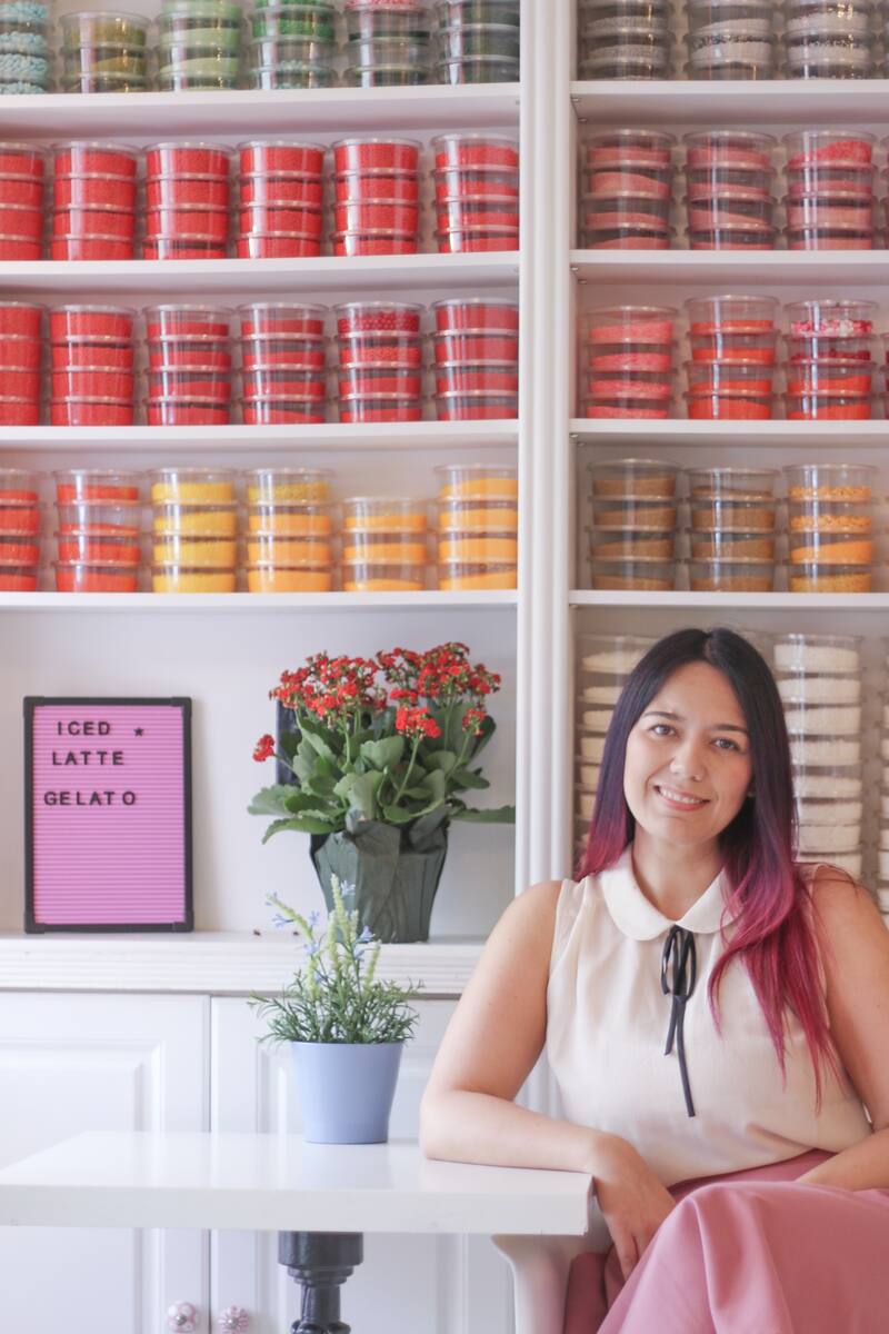 A woman with pink ombre hair sitting at a white table in front of shelves lined with colorful tubs of sprinkles