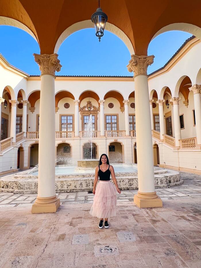 A woman in a pink tulle skirt standing in a courtyard with arches and a fountain