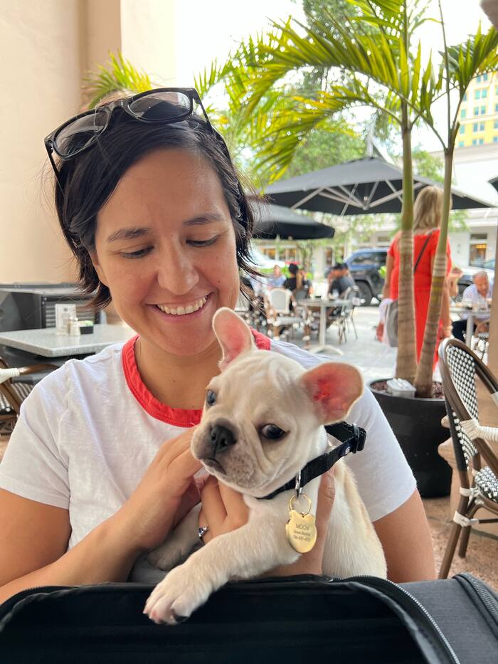 A smiling woman holding a cream French bulldog puppy named Mochi at an outdoor cafe